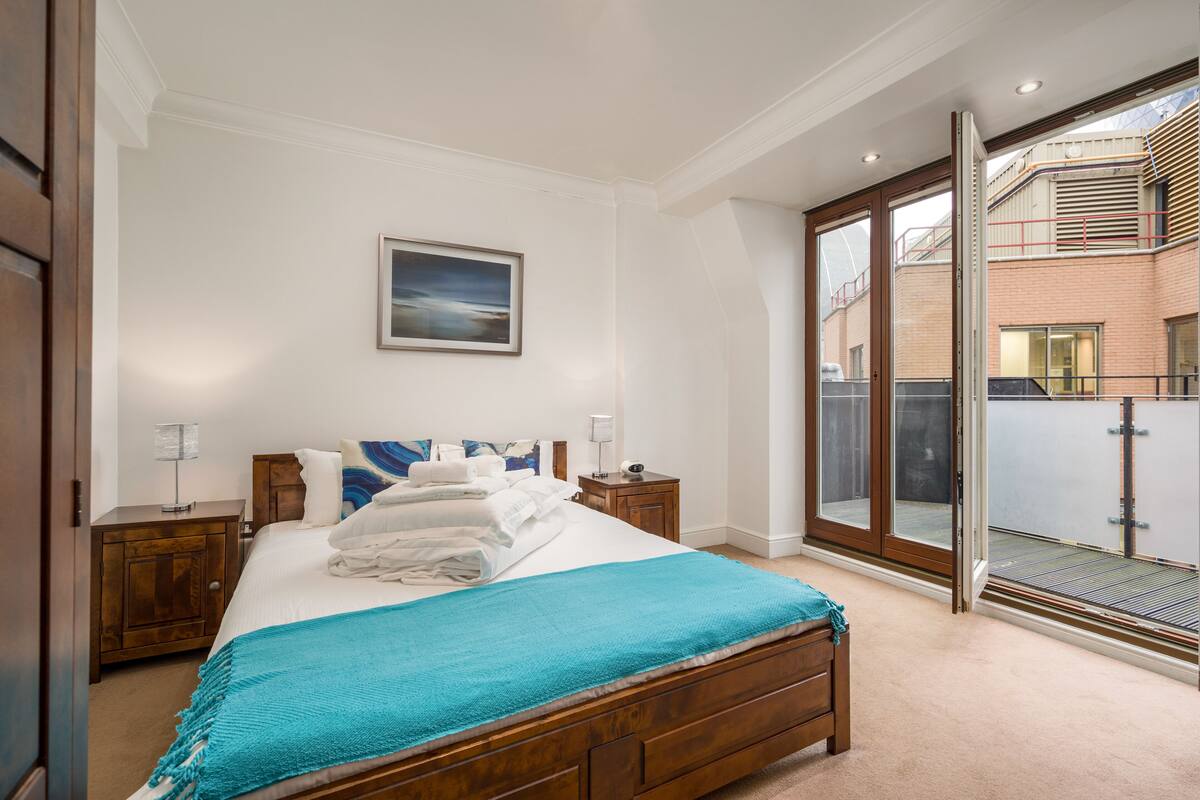A bedroom with a large bed, a nightstand, a bedside table, and a window with a view of a building.