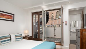 Une chambre à coucher comprenant un lit, une table de chevet, une lampe, une armoire et une vue sur un bâtiment en briques, visible à travers une porte vitrée.
