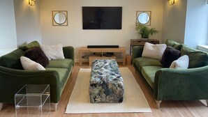 Ett modernt vardagsrum med två gröna soffor, en central pall, en platt-TV och väggmonterade lampor.
