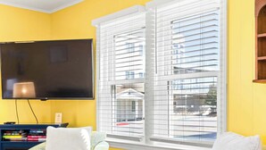Una sala de estar con una pared amarilla, un televisor de pantalla plana, dos sillones estampados, una mesita de madera y una ventana con persianas.