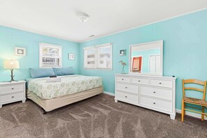 Une chambre à coucher comprenant un lit, une commode, une chaise et deux fenêtres équipées de stores.