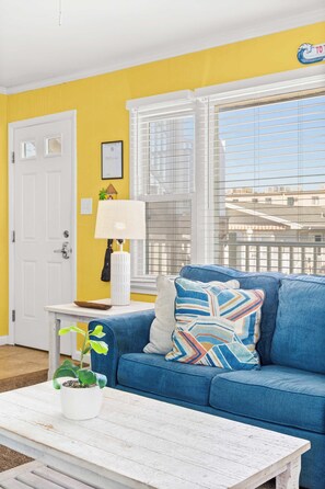 A room with a blue sofa, a white door, a window with blinds, a lamp, and a small plant on a white table.