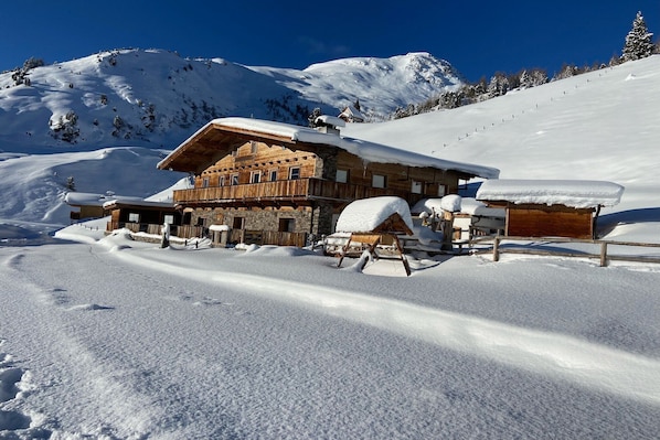 Un chalet en bois, dont le toit est recouvert de neige, situé sur une pente enneigée sous un ciel bleu dégagé.