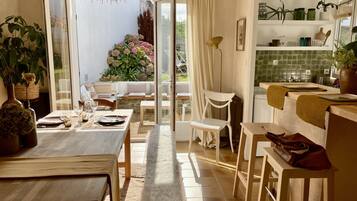 Une salle à manger lumineuse, avec une table dressée pour deux personnes, un banc et une vue sur un jardin, visible à travers une porte vitrée.