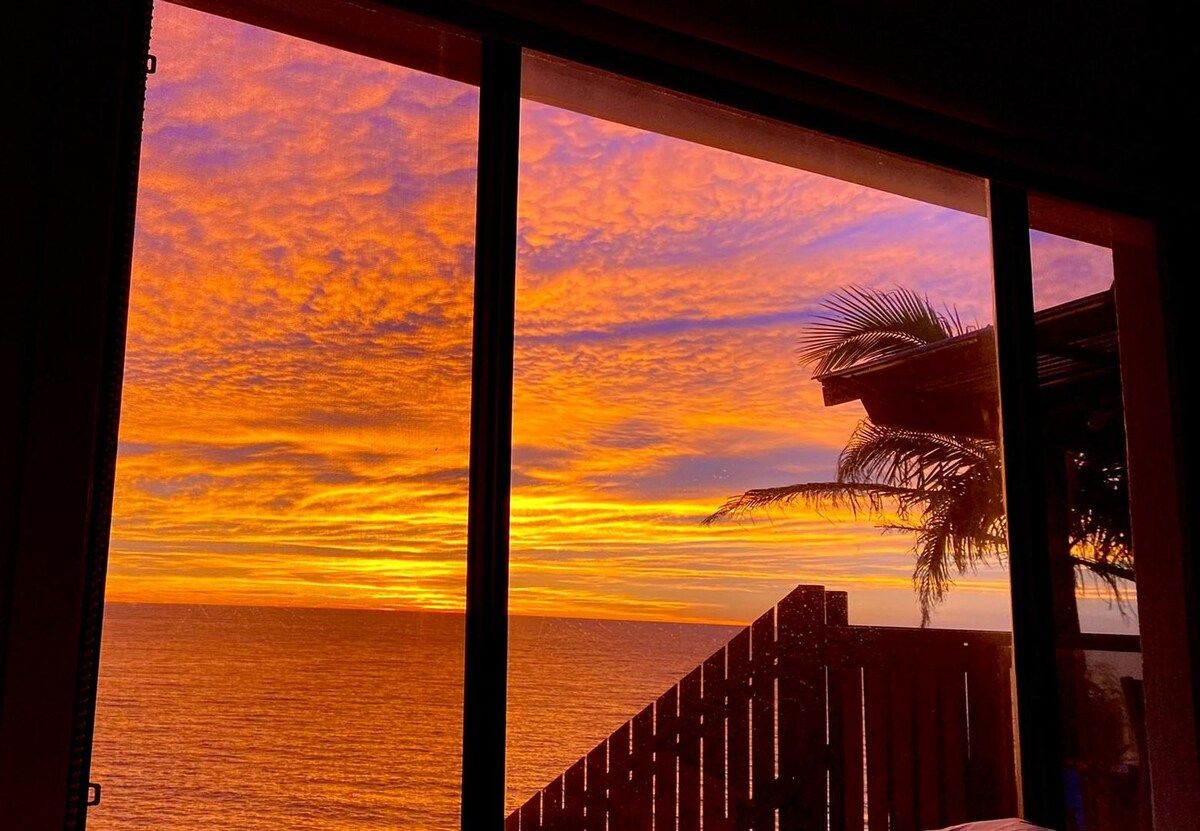 A sunset view through a window with a wooden railing and a palm tree.