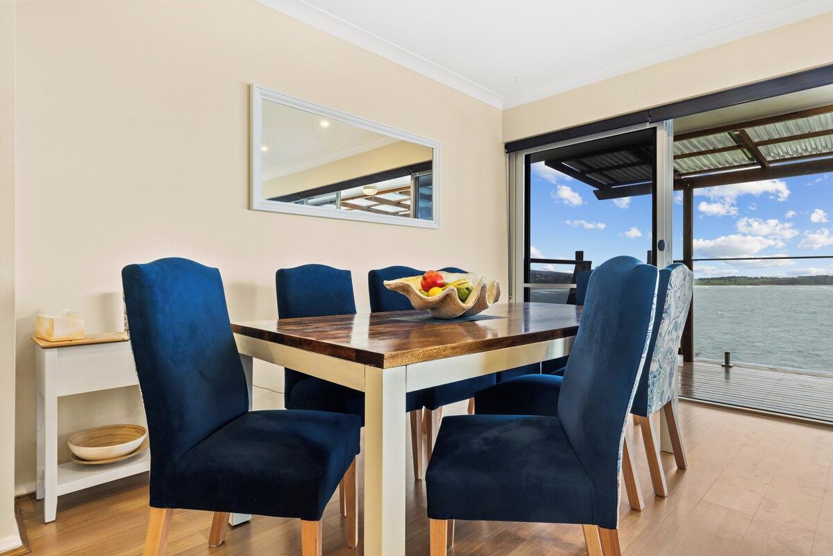 A dining area with a table and chairs, a bowl of fruit, and a view of the water.