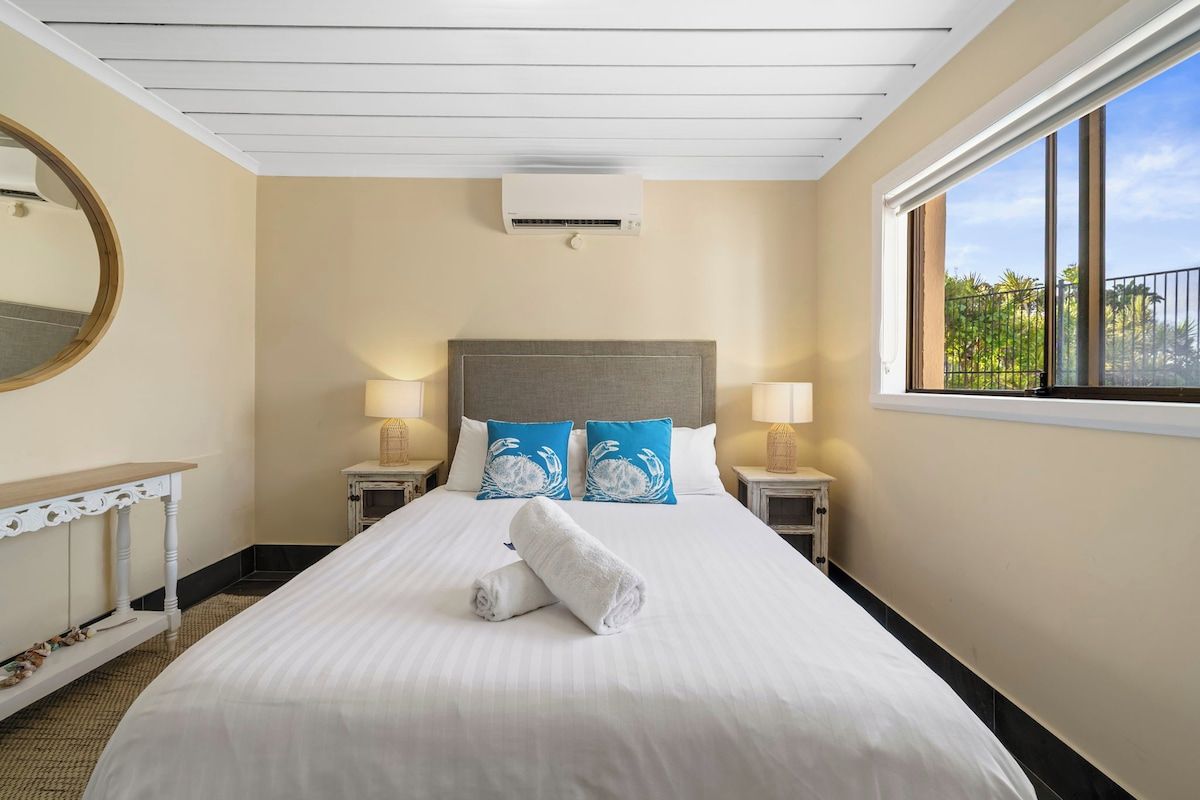 A hotel room with a large bed, two bedside tables with lamps, a round mirror, and a window with a view of greenery.