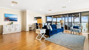 A spacious living room with a blue sofa, a white side table, and a large window offering a view of the sea. - MAGICAL VIEWS OF DEE WHY (Dee Why)