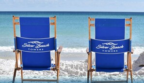 Two blue beach chairs with "Silver Beach Towers" branding, positioned on a sandy beach with ocean waves in the background.
