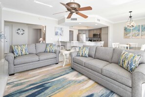 A spacious living room with grey sofas, a colorful striped rug, and a ceiling fan.