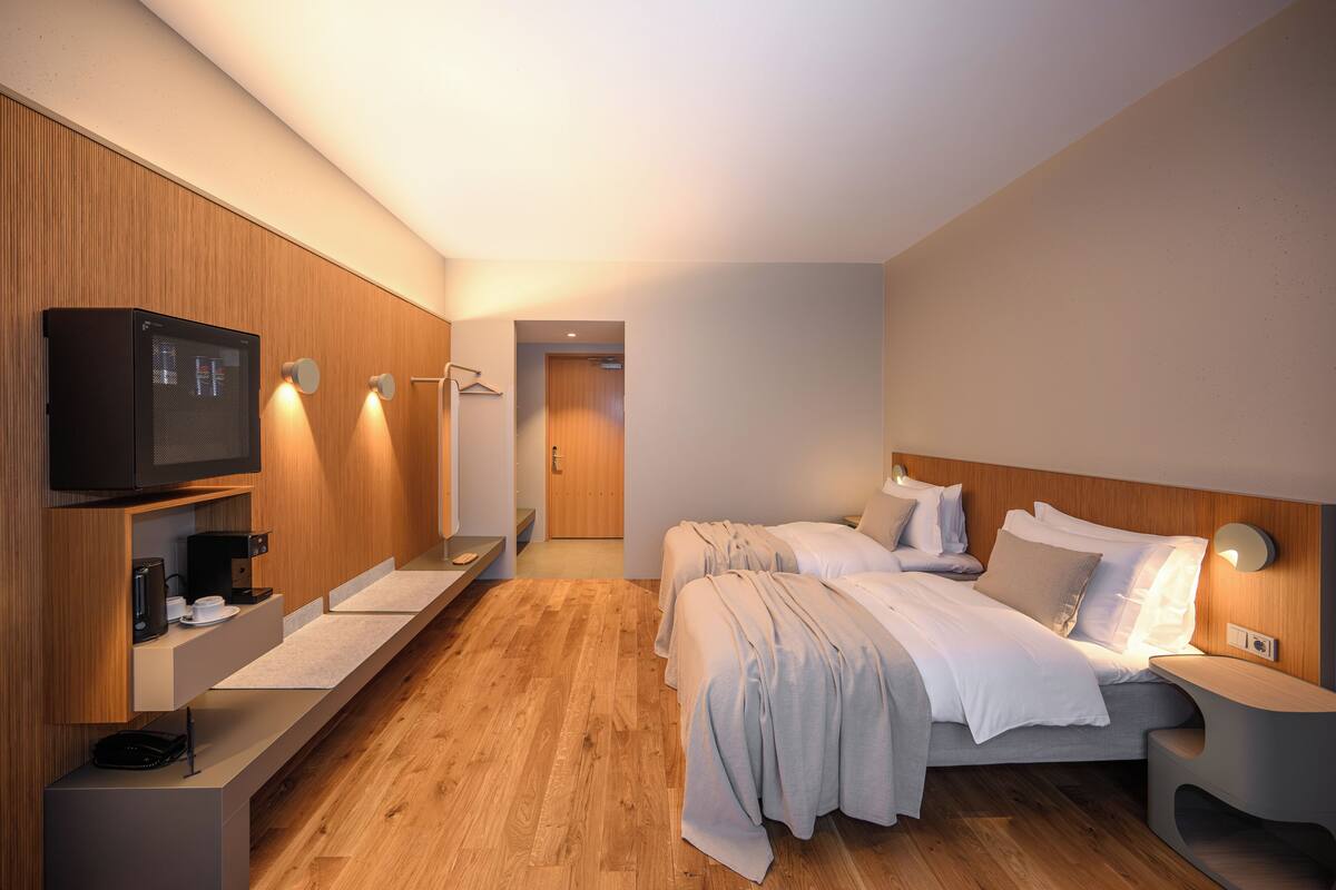 A modern hotel room with a wooden floor, a bed with white linens, a bedside table with a lamp, a TV, and a small kitchenette area.