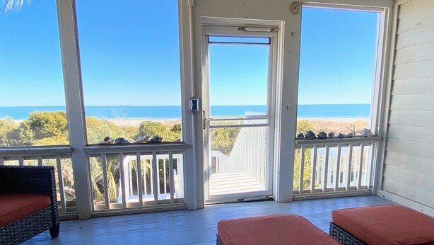 A covered outdoor seating area with wicker furniture and red cushions, overlooking a beach. - Beach Club Villa 29 by Deserving Vacations (Isle of Palms)