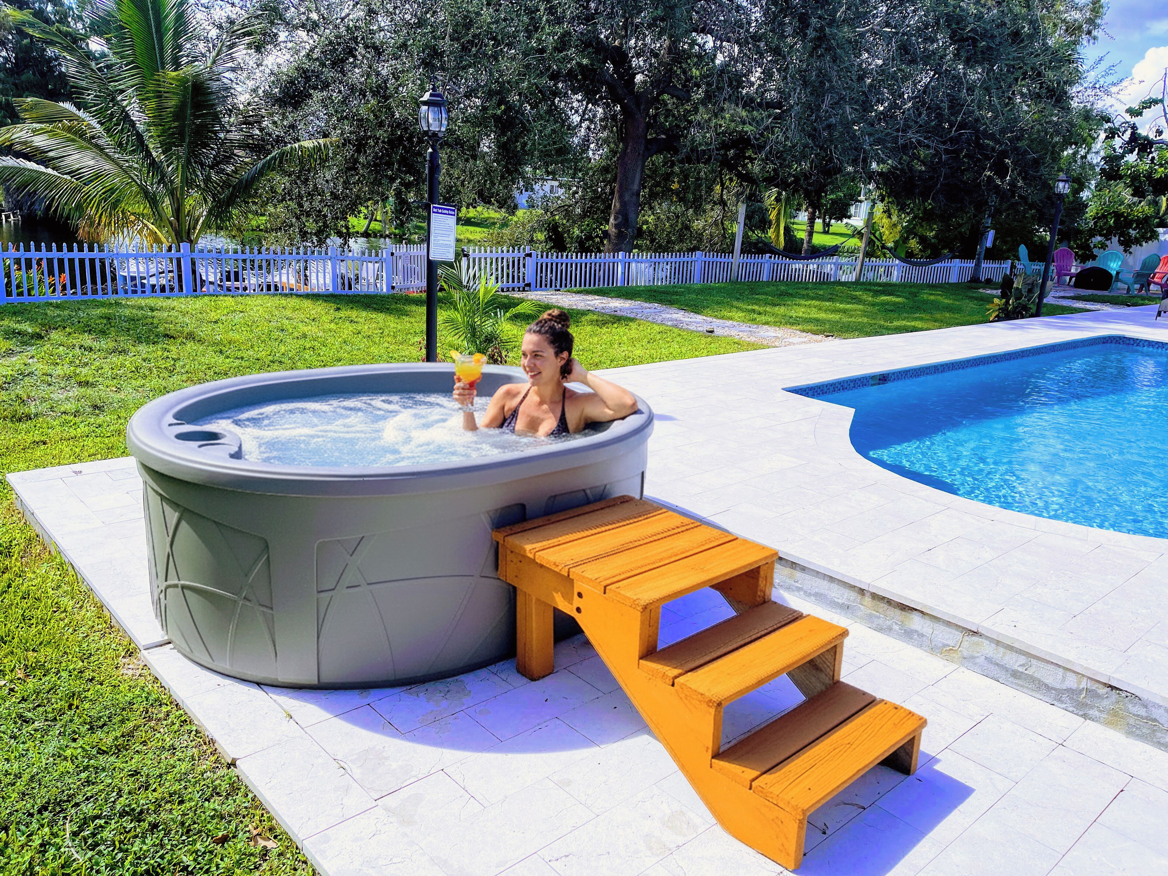 Un jacuzzi con un banco de madera y escalones que lo llevan a él, rodeado por una piscina y una valla blanca.