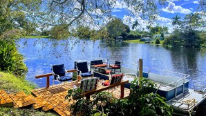 A dock with chairs and tables by a calm lake, surrounded by lush greenery and palm trees.