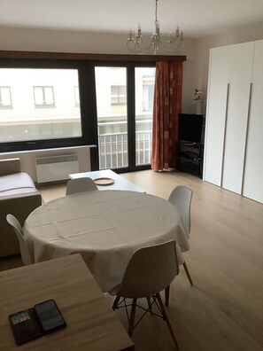 Ein Essbereich mit einem runden Tisch, Stühlen, einer Couch, einem Fernseher und Blick auf ein Gebäude durch ein Fenster.
