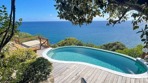 A wooden deck with a swimming pool, surrounded by greenery and overlooking a clear blue ocean.