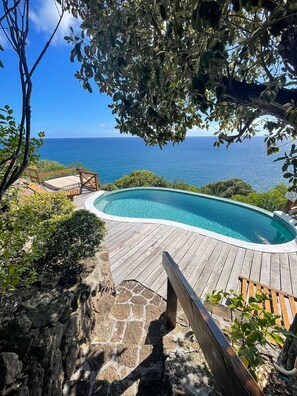 A wooden deck with a swimming pool, surrounded by greenery and overlooking a clear blue ocean.