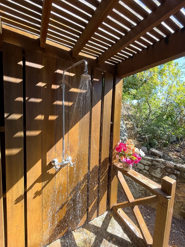A wooden outdoor shower with a metal faucet, surrounded by a wooden pergola and a stone wall.
