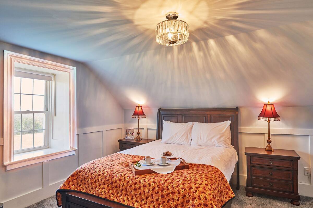 Ein ordentlich bezogenes Bett mit floralem Überwurf, zwei Nachttische mit Lampen, ein Fenster mit Jalousien und ein Chandelier.