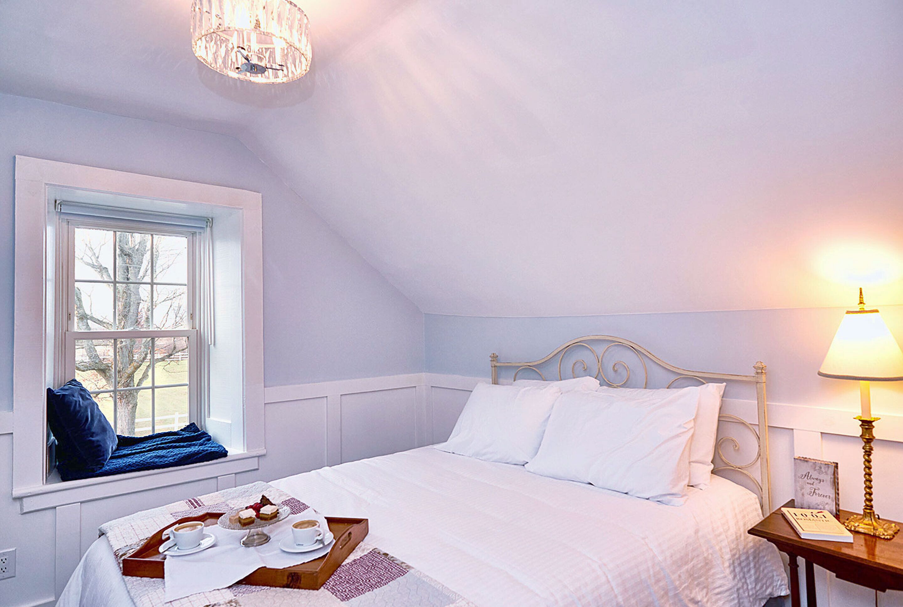 A bedroom with a bed, a window with a view, a bedside table with a lamp, and a tray with breakfast items.