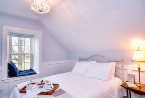 Un dormitorio con cama, ventana con vistas, mesita de noche con lámpara y bandeja con alimentos para el desayuno.