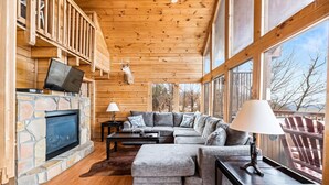 A cozy cabin interior with a stone fireplace, a gray sectional sofa, and a television mounted on the wall. - Charming & Quiet Mtn-Top Cabin w Grand Panoramic Mountain Views!! DOG Friendly! (Sevierville)
