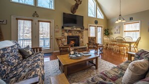 A spacious living room with a stone fireplace, a mounted deer head, and a large TV. There are multiple windows and a dining area with wooden chairs. - 4BDRM Games Family Friendly Fire Pit Masthope (Lackawaxen)