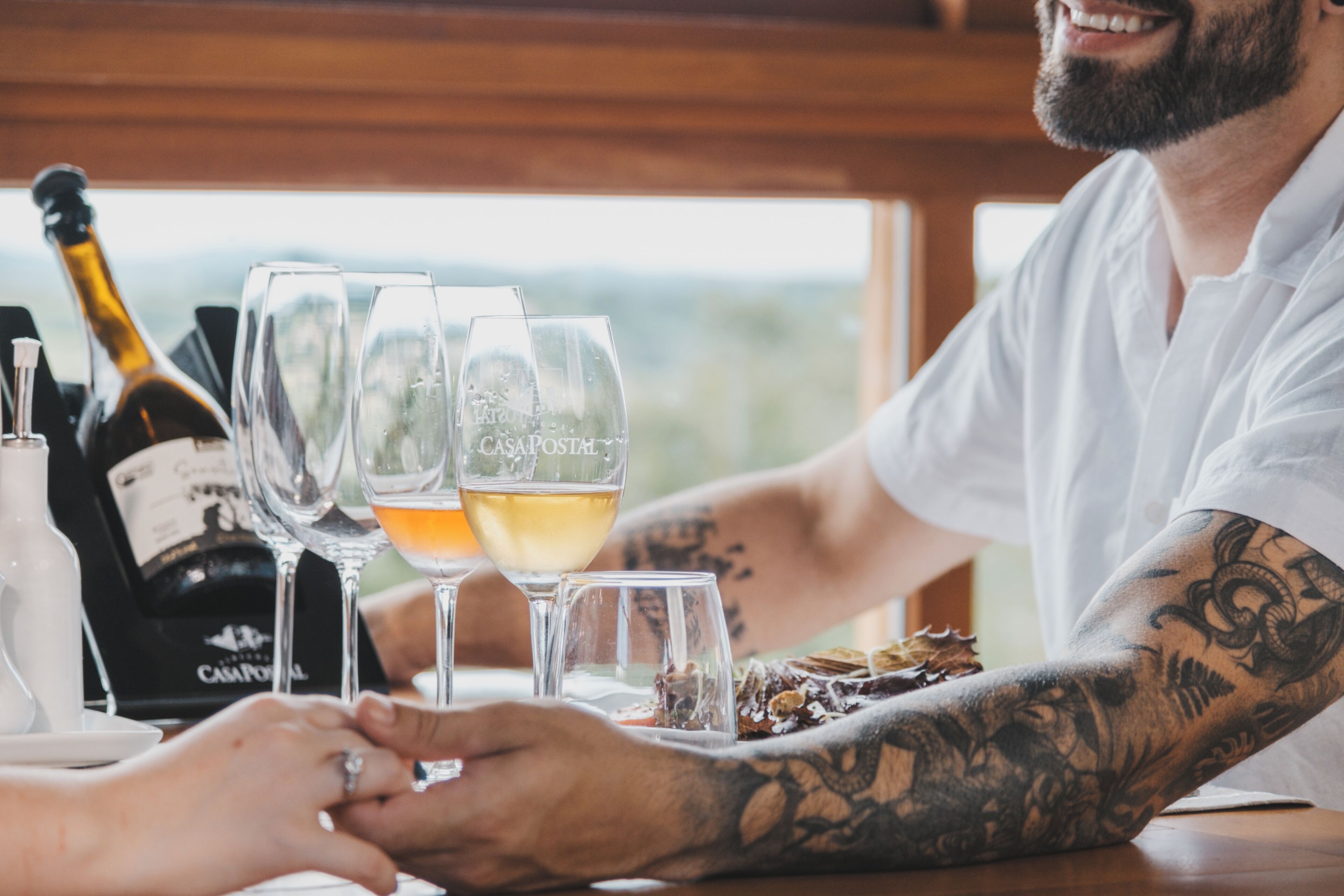 A person with tattoos at a table with wine glasses and a bottle of wine.