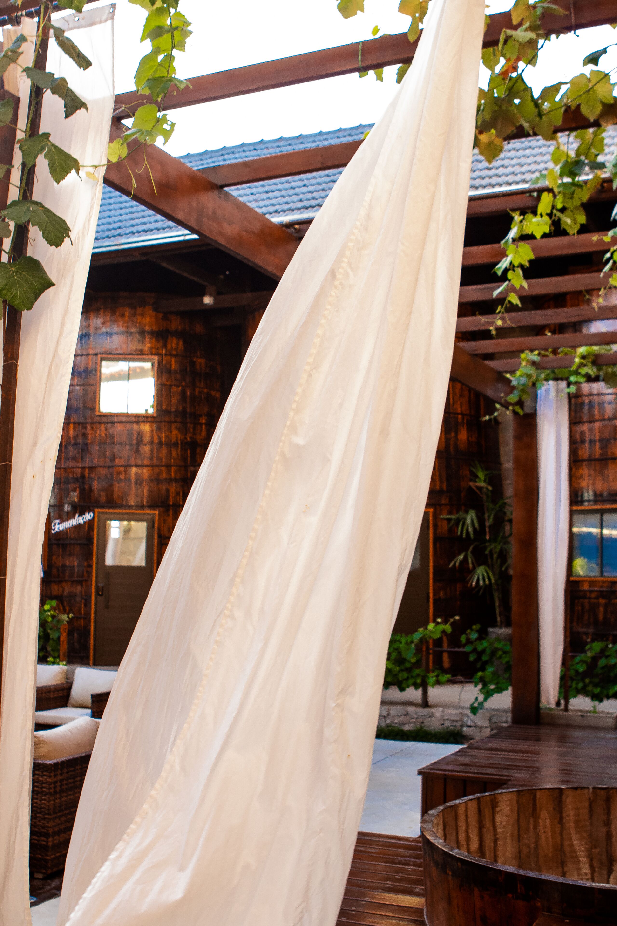 A wooden pergola with white curtains, a wicker sofa, and a wooden table.
