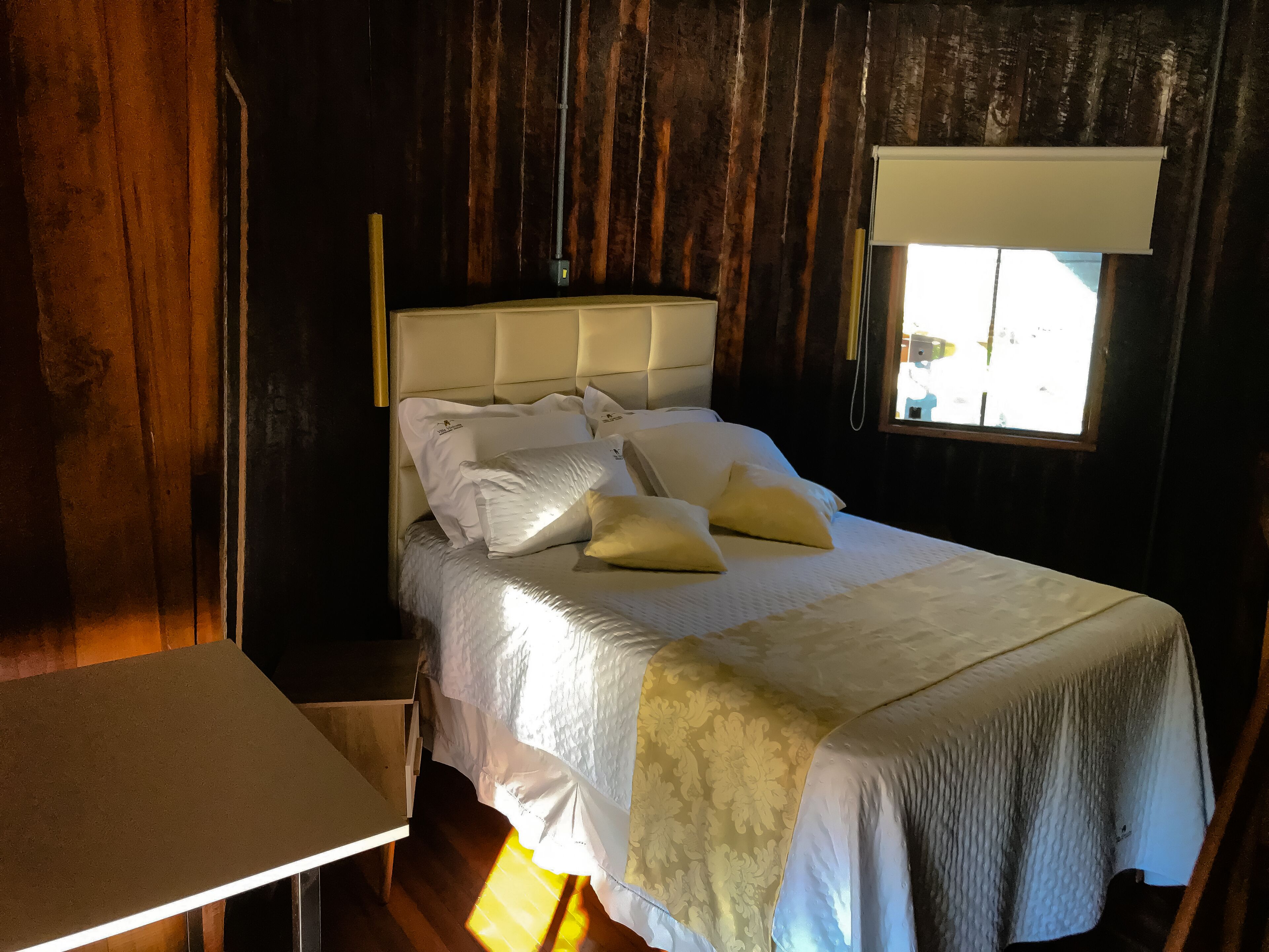 Una cama con cabecera, almohadas y mesita de noche en una habitación con paredes de madera y una ventana.