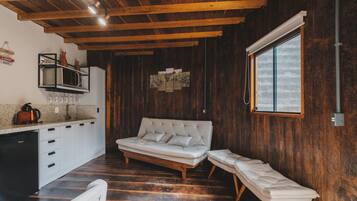 Um quarto acolhedor com teto de madeira, um sofá, um banco, uma cozinha com geladeira e uma janela que oferece luz natural.