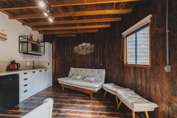A cozy room with a wooden ceiling, a sofa, a bench, a kitchen area with a refrigerator, and a window providing natural light.