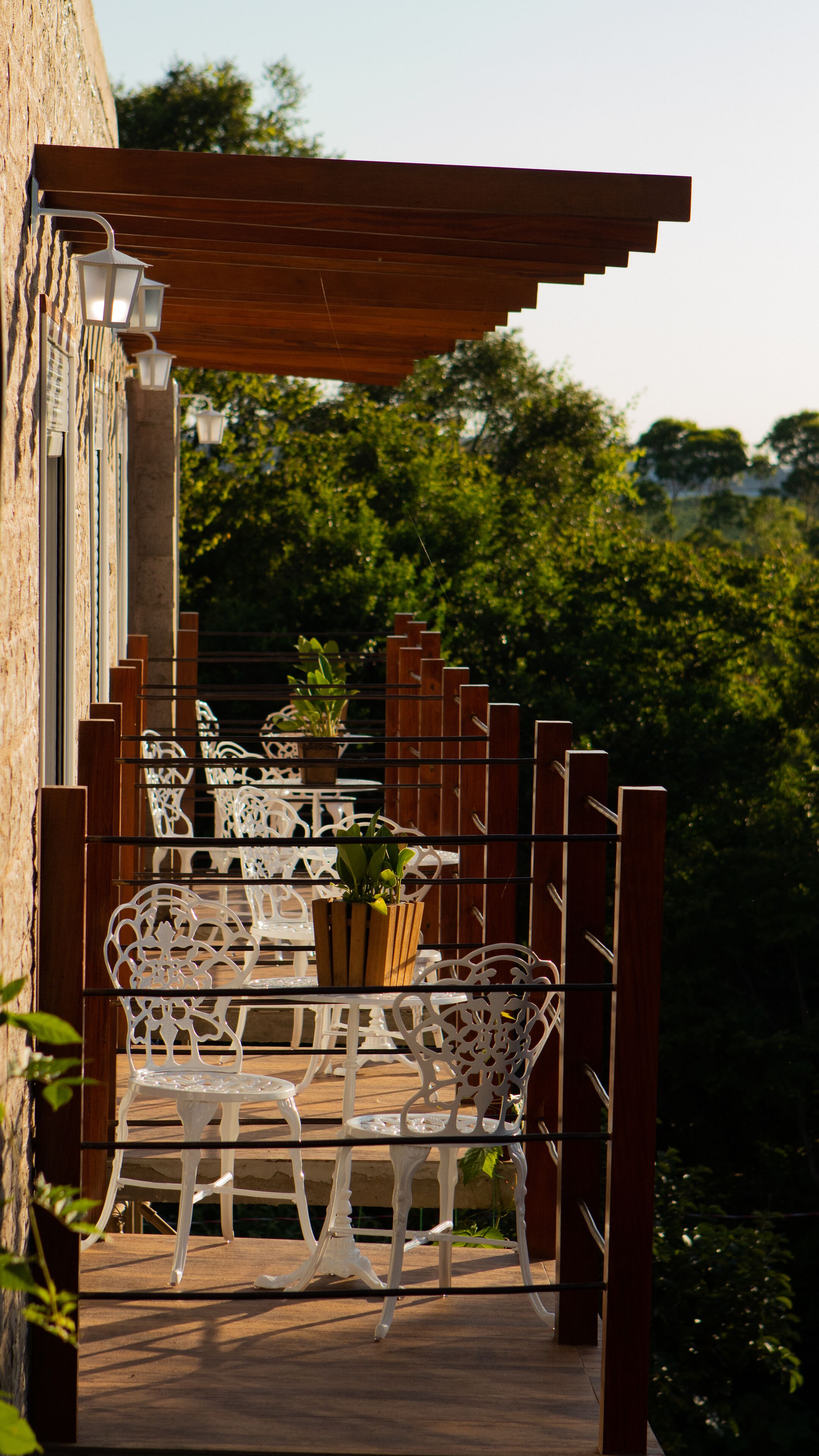Una pérgola de madera con sillas y mesas de metal blanco, rodeada de follaje.