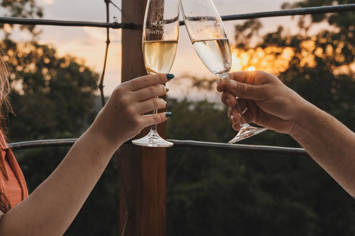 Two people toasting with champagne glasses at a round table with a decorative metal top, outdoors with a sunset in the background.