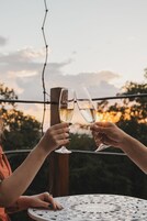 Duas pessoas brindando com taças de champanhe em uma mesa redonda com tampo de metal decorativo, ao ar livre com o pôr do sol ao fundo.