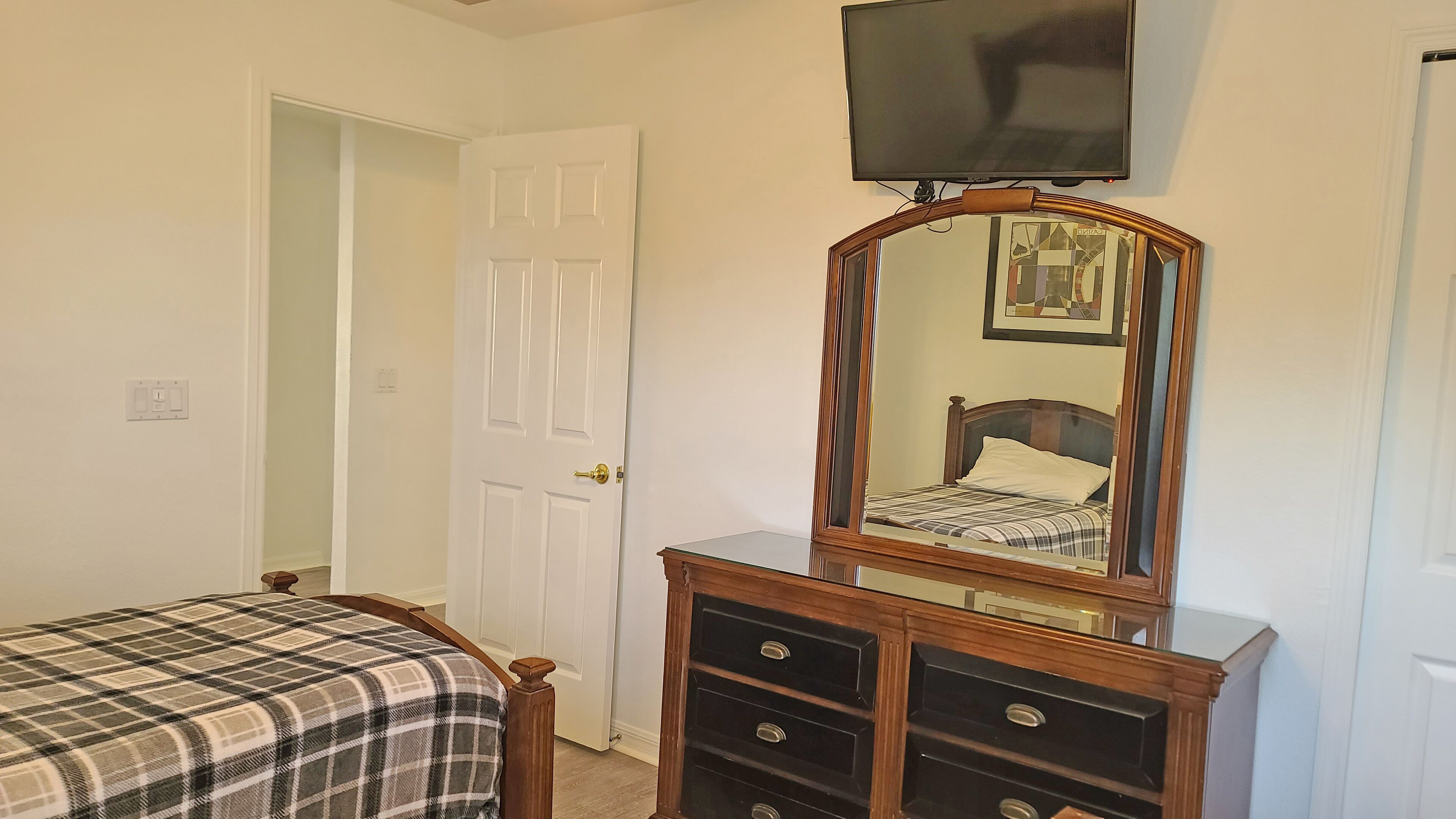 Un dormitorio con una cama, un aparador con cajones, un espejo y un televisor montado en la pared.