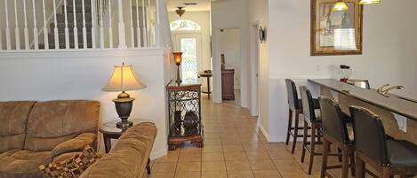 A spacious living room with a staircase, a dining area with bar seating, and a kitchen with a large window.
