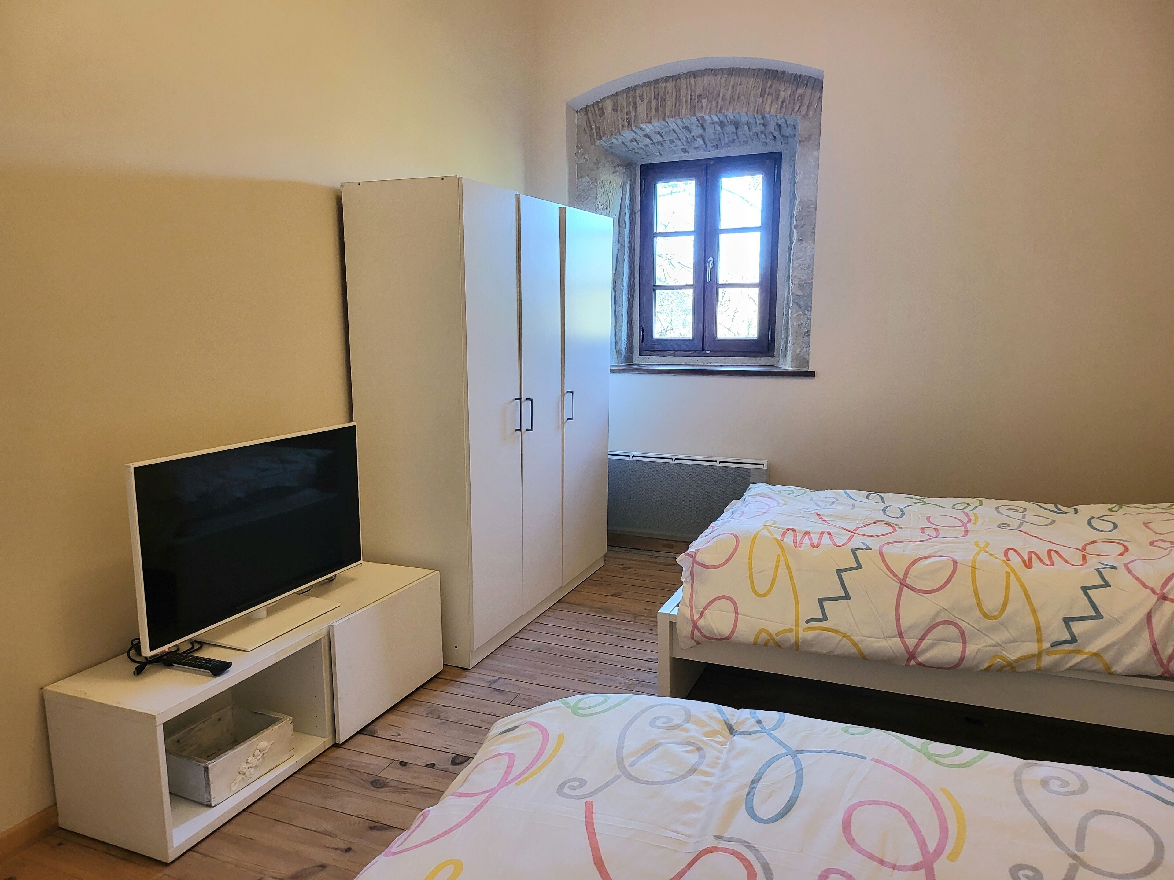 A bedroom with a bed, a TV on a stand, a wardrobe, a window, and a radiator.