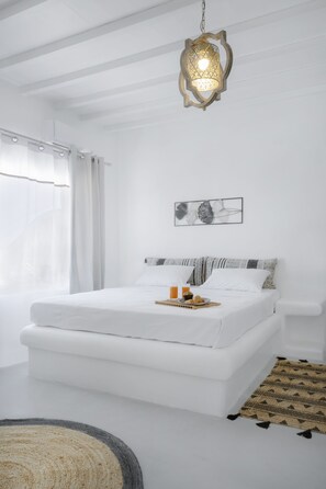 A white bedroom with a bed, a hanging light fixture, a window with sheer curtains, and a framed picture on the wall.