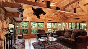 A rustic lodge interior with a stone wall, wooden ceiling beams, a large window, a fireplace, and a leather sofa. - On the Eagle River Chain O’ Lakes, Lake Front, 3200 sq. ft., 2.5 baths (Eagle River)
