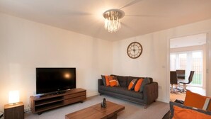 A modern living room with a flat-screen TV, a sofa with orange and gray cushions, a wooden coffee table, and a chandelier. - The Terrace (Derby)
