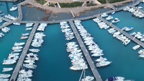 Uma marina com vários barcos atracados, uma estrutura semelhante a um castelo numa colina, e edifícios com telhados vermelhos.