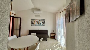 Uma sala de estar com uma mesa branca, cadeiras brancas, um sofá escuro, um quadro de barcos à vela e uma janela com cortinas leves.