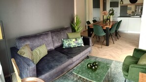 Una sala de estar moderna con un sofá gris oscuro, cojines verdes, una mesa de centro de vidrio y una zona de comedor con mesa de madera y sillas verdes.