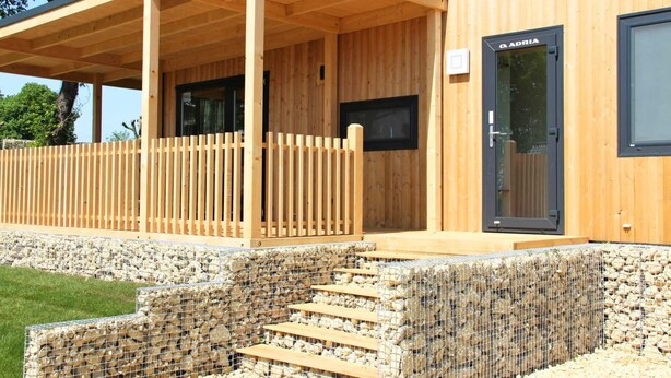 Un edificio de madera con un porche cubierto, una puerta con el letrero "ADRIA" y una escalera que lleva a un muro de piedra.