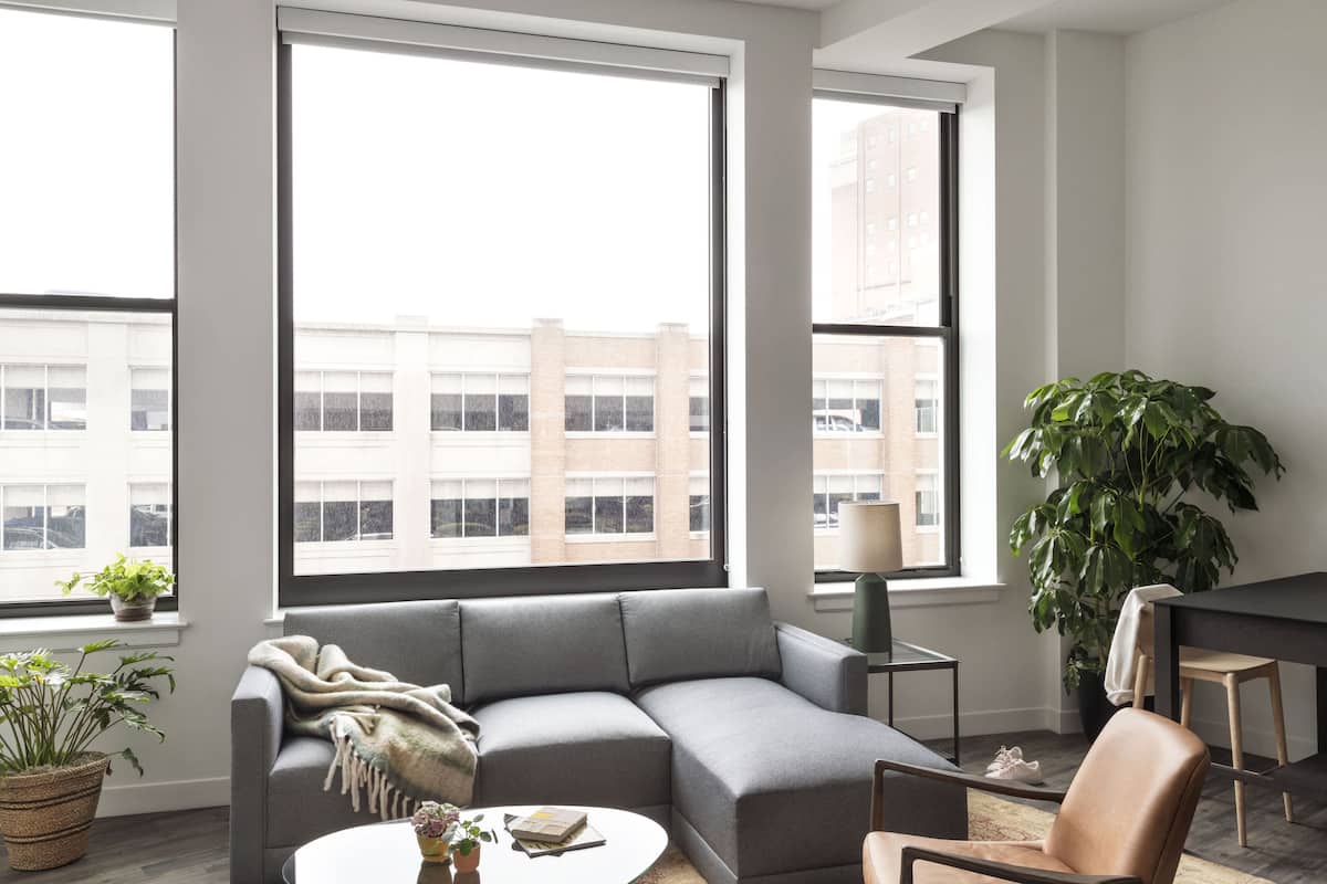 A modern living room with a grey sectional sofa, a round coffee table, and a large window offering a view of a building.