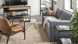 A modern living room with a flat-screen TV, a sofa, a coffee table, and a chair. There is a large window providing natural light and a view of the outdoors. - ROOST Cleveland (Cleveland)