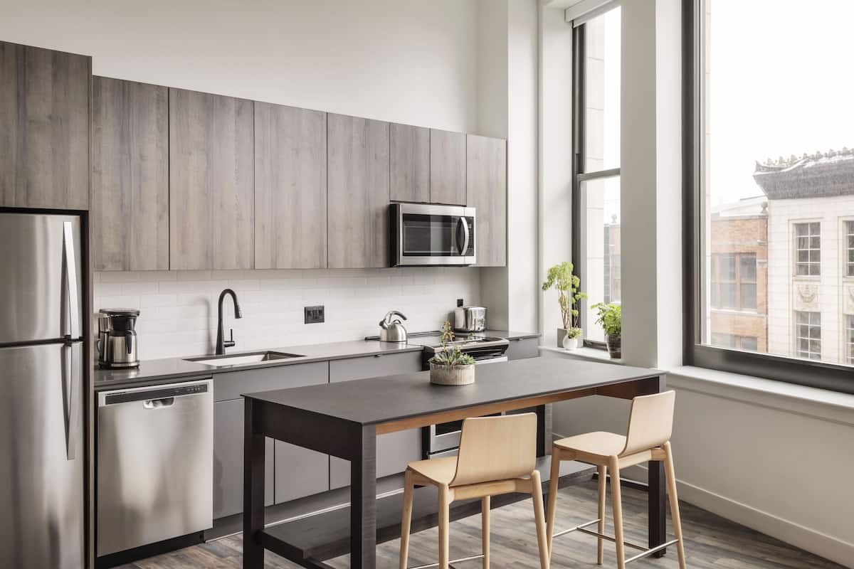 A modern kitchen with grey cabinets, stainless steel appliances, a dark countertop, and two wooden chairs.