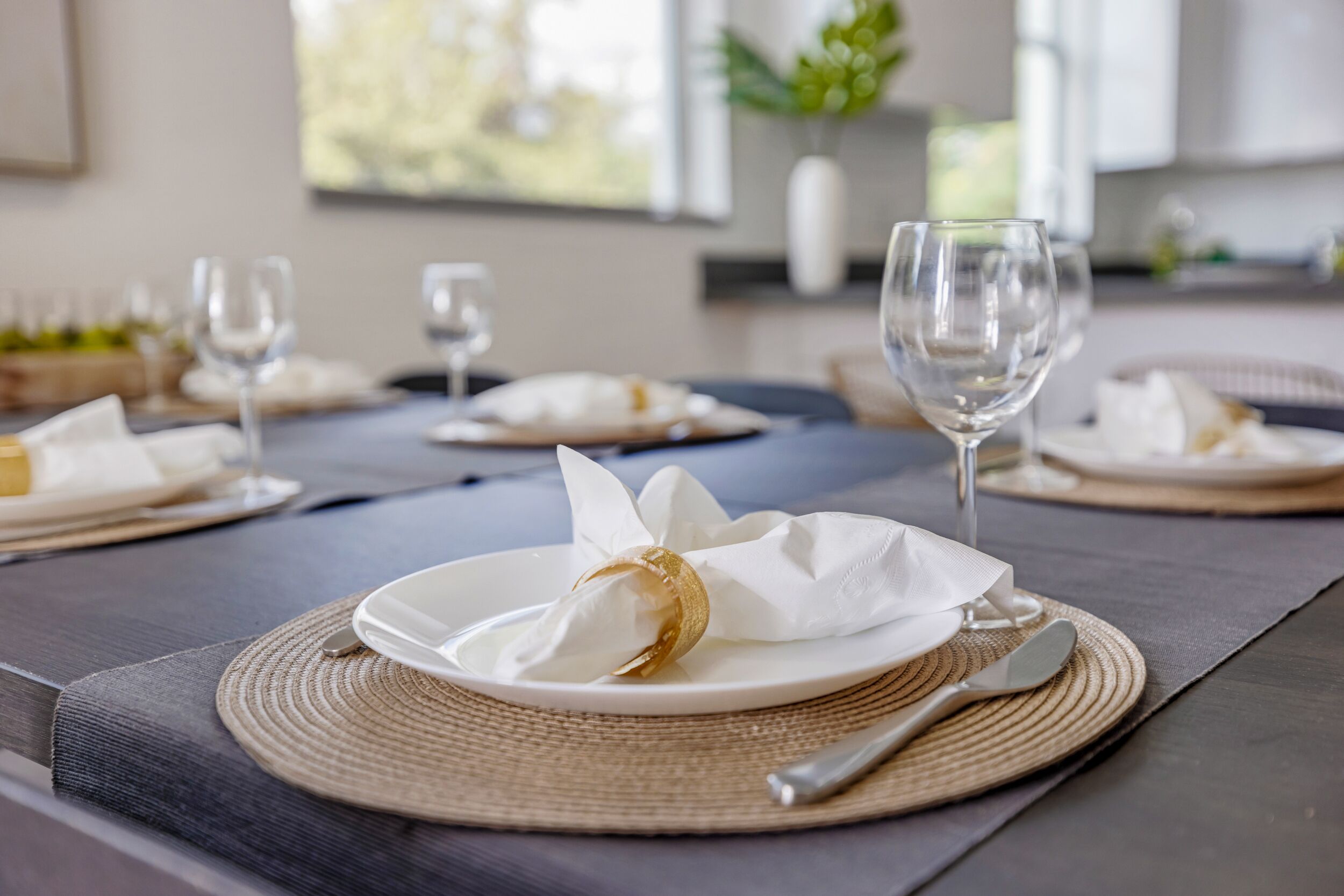 A dining table set with white plates, silverware, and wine glasses, with a woven placemat and a decorative napkin centerpiece.