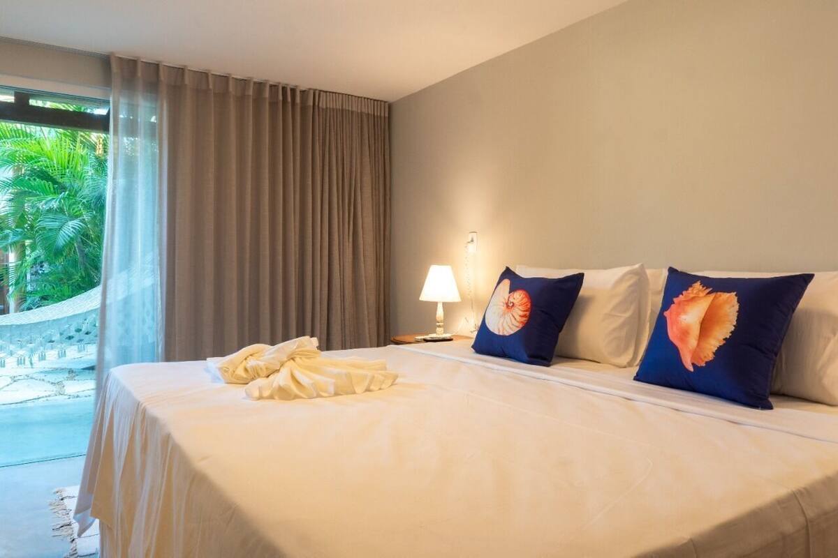 Quarto de hotel com cama, cortinas, luminária e vista para área verde lá fora.